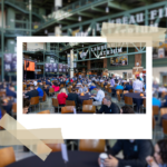 A large crowd sits at round tables inside the Lambeau Field Atrium, surrounded by brick walls, green steel beams, and banners honoring retired Green Bay Packers players. A big screen and “Lambeau Atrium” signage hang above the event space, creating a lively, stadium‑themed atmosphere.